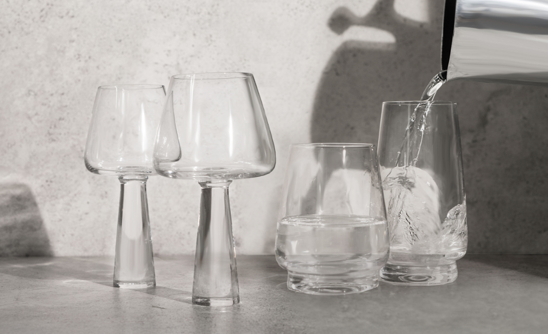 Modern glassware collection with various shapes, including water being poured into a glass, set against a concrete surface.