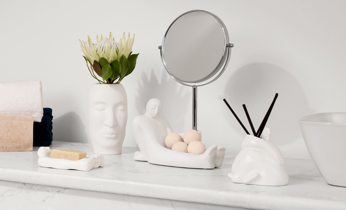 Decorative white ceramic vases and sculptures with face designs, surrounded by candles and greenery on a dark surface.