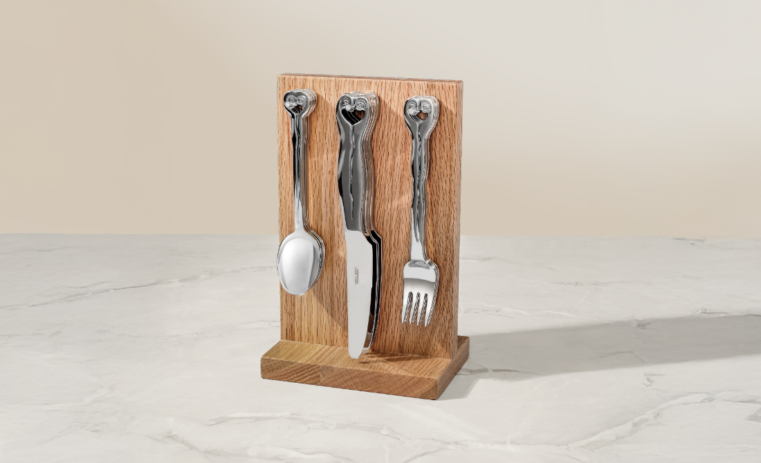 Polished silverware with wave-themed handles displayed on a wooden stand, alongside a stainless steel breadbox and a loaf of bread.