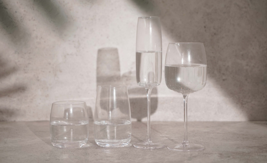 Assorted glassware including wine glasses and tumblers, filled with water and beverages, casting shadows on a surface.