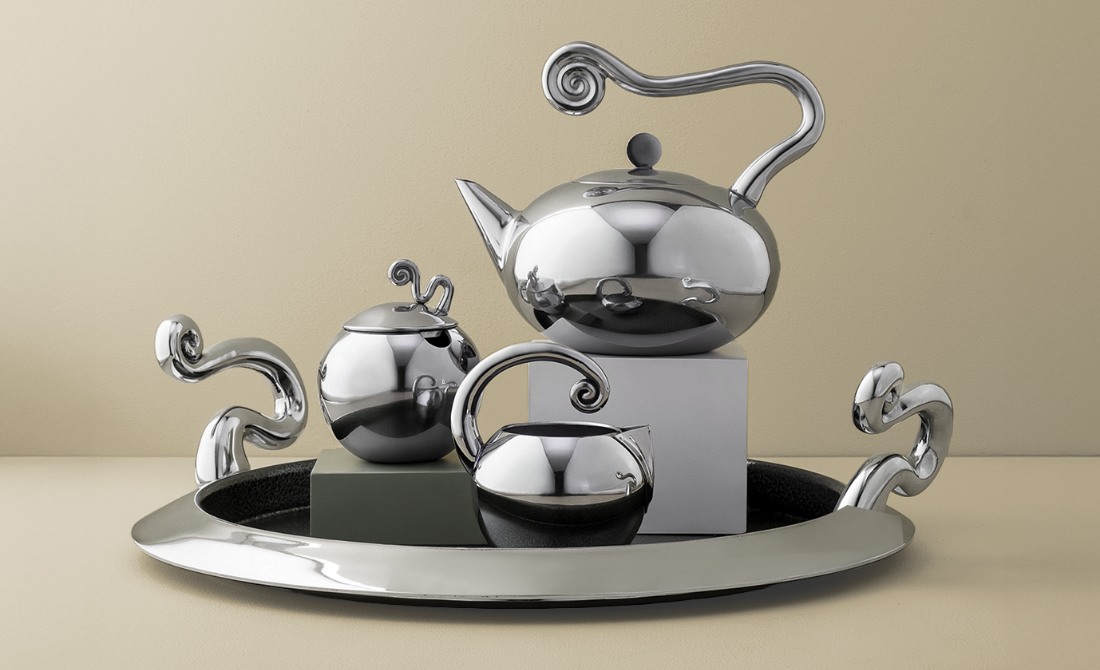Polished metallic tea set with wavy handles, including a teapot, sugar bowl, and creamer, arranged on a tray against a blue background.